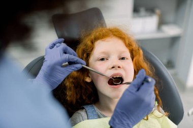Diş kliniğinde bir çocuk. Kırmızı kıvırcık saçlı küçük bir kızın portresi, dişçi koltuğunda ağzı açık bir şekilde otururken dişi Afrikalı dişçi diş aynası kullanarak diş kontrolü yapıyor.