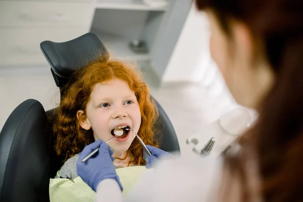 Visão traseira superior do dentista feminino em luvas de látex azul, verificando a condição dos dentes de uma menina encaracolada vermelha sorridente, sentada na cadeira dentária na clínica odontológica moderna. Odontologia pediátrica — Fotografia de Stock