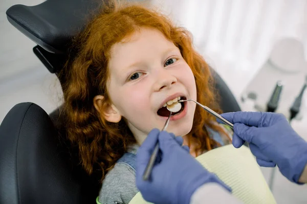 Dziecko na przyjęciu u dentysty. Close up portret ślicznej dziewczynki z czerwonymi kręconymi włosami, siedzi z otwartymi ustami w fotelu stomatologicznym, podczas gdy dentysta sprawia, że zęby sprawdzić za pomocą lusterka dentystycznego — Zdjęcie stockowe