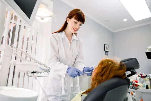 Side view of happy pretty doctor in white uniform and gloves, making examination or curing caries and toothache for her little patient, red haired curly girl, sitting in dental chair — Stock Photo, Image