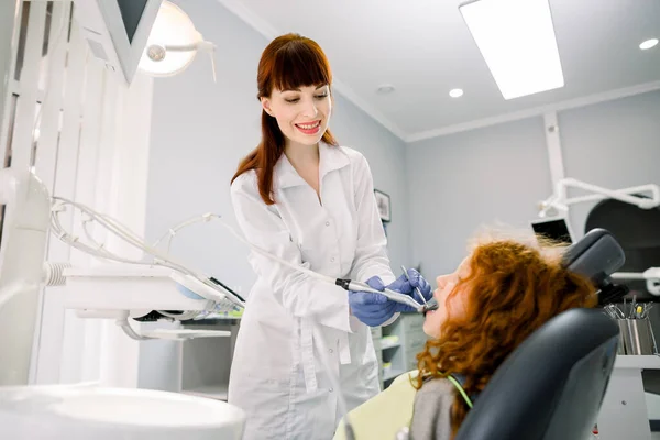 Shot of young pretty female Caucasian dentist in white uniform, making dental check up and caries treatment with drill for her cute little patient, red haired girl, sitting in dental chair at clinic —  Fotos de Stock