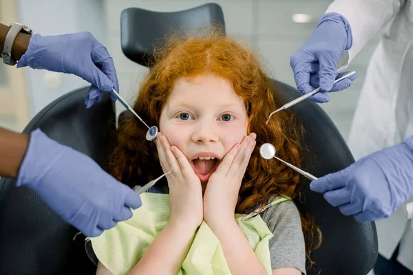 Healthy teeth, caries prevention and pediatric dentistry. Close up of scared funny little red haired curly girl, looking at camera and screaming and hands of two dentists in gloves with dental tools