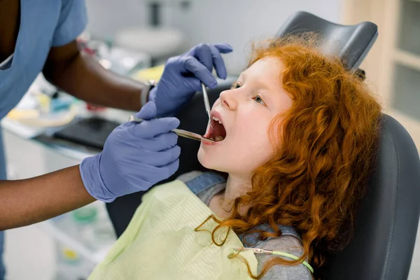 Zbliżenie małe słodkie kręcone rude włosy dziewczyna z otwartymi ustami podczas badania doustnego u dentysty. Ręce afrykańskiego dentysty w lateksowych rękawiczkach, robiącego badania zębów narzędziami — Zdjęcie stockowe