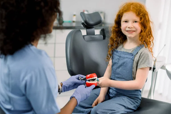 Szczęśliwy uśmiechnięty dzieciak w gabinecie dentystycznym. Mała dziewczynka z czerwonymi kręconymi włosami, siedzi na fotelu dentystycznym i patrzy w kamerę, podczas gdy jej afrykańska dentystka pokazuje jak prawidłowo myć zęby na modelu szczęki — Zdjęcie stockowe