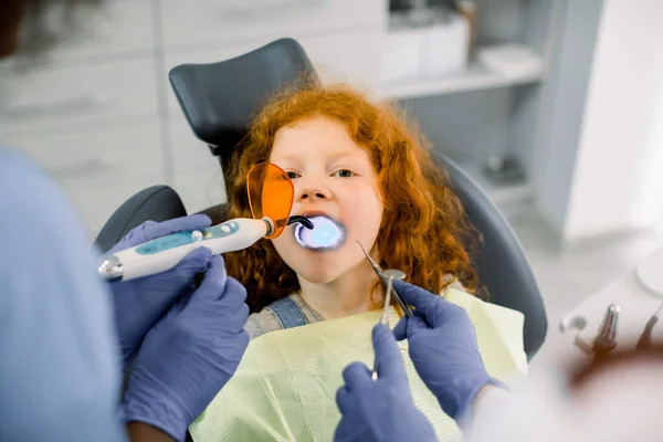 Visão de perto da menina paciente fazendo tratamento odontológico no consultório de dentistas. Mãos de duas dentistas femininas que fazem restauração dentária com enchimento usando polimerização de cura lâmpada UV — Fotografia de Stock
