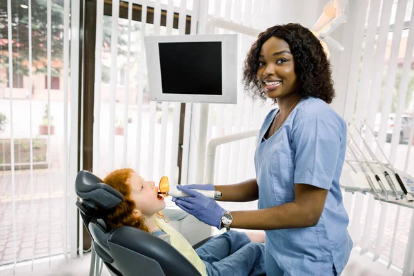 Medicina, stomatologia e concetto di cura dei denti. Giovane sorridente dentista donna africana con lampada UV cura dentale, guardando la fotocamera, finitura riparazione dei denti per la sua piccola paziente, carina ragazza — Foto Stock