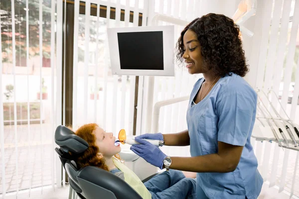 Female African dentist performing dental filling procedure to a little red haired girl in pediatric dental clinic. Doctor using dental ultraviolet curing light for filling polymerization