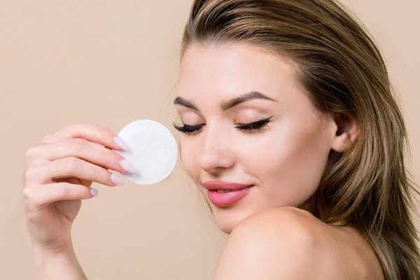 Young pretty Caucasian blond woman cleaning her face, using cotton pad with cleansing lotion or facial toner for removing makeup. Skin care concept. Beige background, copy space —  Fotos de Stock