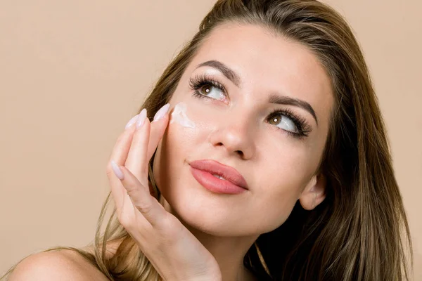 Close up beauty portrait of beautiful naked Caucasian blond woman, applying moisturizing cream on her flawless skin and looking up, posing on isolated beige background. Skin care, face cream —  Fotos de Stock