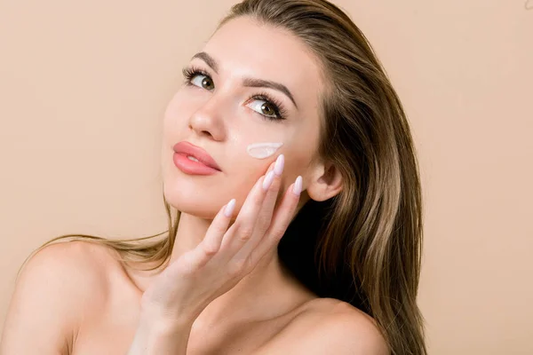 Young European blond woman with moisturizing cream on cheek. Beauty shot of beautiful woman touching her fresh glowing hydrated facial skin on beige background. Youth and Skin care concept —  Fotos de Stock