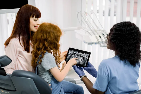 Young African woman dentist showing dental x-ray on tablet to his little red haired girl patient and her mother before medical procedure in clinics. Pediatric dentistry and orthodontics