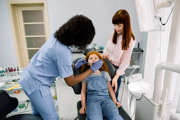 Jovem dentista afro-americana com ferramentas dentárias, fazendo check-up dental de sua menina paciente. Jovem mãe tranquilizando sua filha fofa assustada durante o exame — Fotografia de Stock