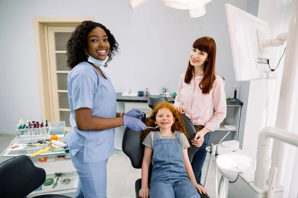 Young smiling mother with her cute preteen daughter, visit their pediatric dentist, African woman, for regular teeth checkup at stomatology clinic. Child with mom at dentists office