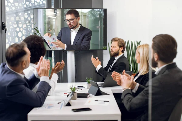 Skupina pěti šikovných multietnických podnikatelů tleská rukama a dívá se na obrazovku, pořádá videokonferenci v zasedací místnosti, kde jejich mužský kolega vysvětluje některé finanční diagramy — Stock fotografie
