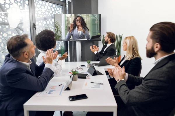 Videokonference, telemeeting, vzdálená práce. Multiethnic různorodý obchodní tým, sedí kolem stolu v kanceláři, pozdravuje své africké kolegyně, tleskání rukou a při pohledu na televizní obrazovku — Stock fotografie
