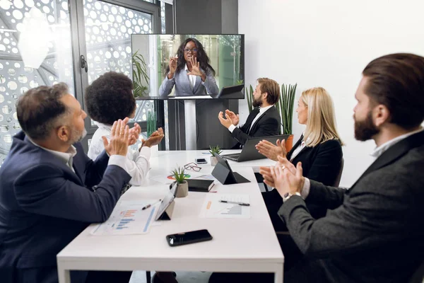 Vidéoconférence, télémarketing, télétravail. Équipe multiethnique d'affaires diverses, assis autour de la table dans la salle de bureau, saluant leur collègue africaine, applaudissant les mains et regardant l'écran de télévision — Photo