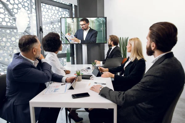 Team af fem fokuserede multietniske forretningsfolk kigger på skærmen, har videokonference i moderne kontor, med deres mandlige kollega forklarer nogle finansielle diagrammer. Telekommunikation - Stock-foto