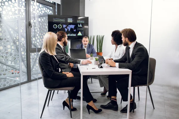 Tým pěti různorodých multietnických podnikatelů, kteří se scházejí v zasedací místnosti v kanceláři v interiéru, sedí u stolu s tabletami a notebookem, před obrovskou plazmovou televizí — Stock fotografie