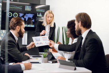 Çekici, kendine güvenen, deneyimli sarışın kadın liderin, odaklanmış profesyonel çok ırklı ortaklarıyla yaptığı toplantıda, gazetede bilgi grafikleri ve finansal tabloları göstermesi.