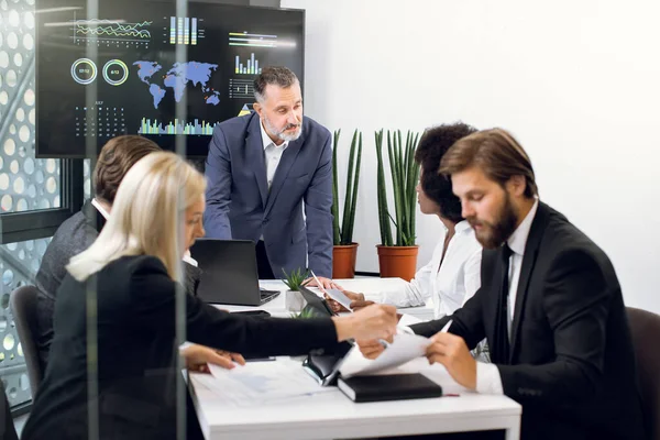 Yetişkin bir erkek patronla, çeşitli etnik çeşitlilikte yöneticilerden oluşan bir ekip konferans odasında görüşlerini paylaşıyor, gazetelerle, tabletlerle ve istatistiklerle büyük bir televizyon ekranının arkasında çalışıyor.
