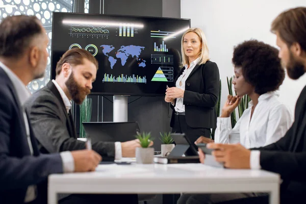 Femme d'affaires blonde agréable et confiante d'âge moyen, montrant des infographies de travail des entreprises sur écran mural numérique pendant la réunion avec l'équipe de divers hommes et femmes d'affaires multiraciaux — Photo