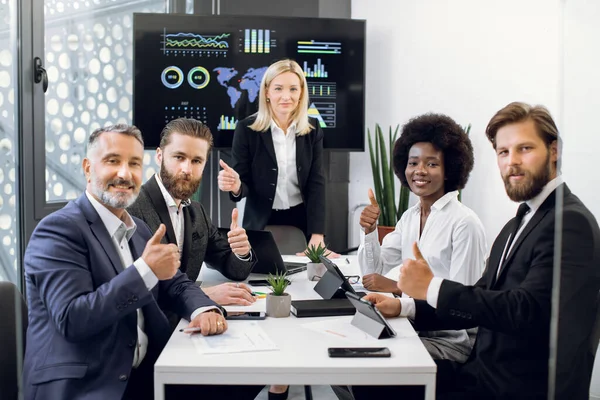 Tým profesionálních vysoce kvalifikovaných multietnický tým obchodních mužů a žen, sedí u stolu v kanceláři s velkou stěnou obrazovky, a ukazuje palce nahoru na kameru, zblízka — Stock fotografie