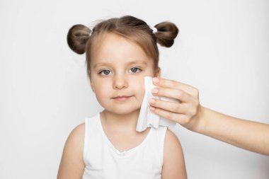 Çocuklar hijyen ve cilt bakımı, ıslak mendil tanıtımı. Anne elinin ıslak mendille bebek yüz derisini silerken yakın plan. Beyaz bluzlu küçük kız, beyaz arka planda kameraya bakıyor.