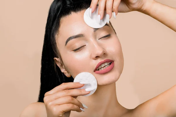 Jovem mulher asiática com os olhos fechados, gostando de limpar o rosto, usando almofadas de algodão com loção de limpeza ou toner facial para remover a maquiagem. Conceito de cuidados com a pele. Fundo bege, espaço de cópia — Fotografia de Stock