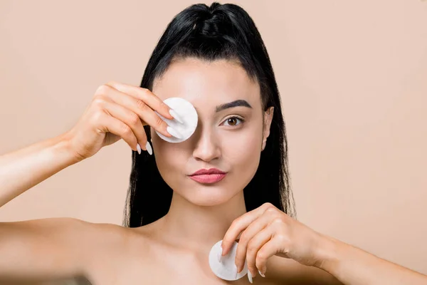 Linda jovem morena asiática segurando almofadas de algodão, escondendo um olho e sorrindo, cuidando de seu rosto para uma pele fresca e saudável, posando contra fundo bege. Cuidados com a pele e beleza — Fotografia de Stock