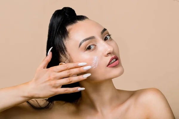 Cuidado com a pele. Linda modelo asiática de morena feminina com pele brilhante e maquiagem natural, aplicando creme em seu rosto, posando para a câmera em fundo bege, close-up. Conceito de hidratação da pele — Fotografia de Stock