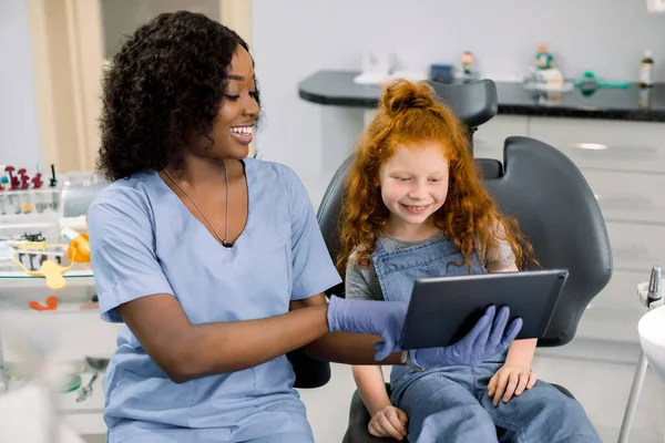 Medicine, pediatric dentistry and oral care concept. Female smiling African dentist showing tablet pc computer to happy kid patient, curly red haired school girl at modern dental clinic