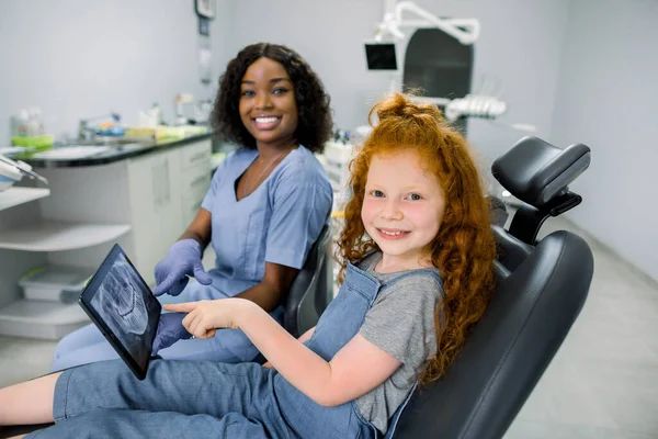 Gyerek a fogorvosi rendelőben. Kis türelmes vörös hajú lány, ül a fogorvosi székben, nézi a kamerát, és rámutatva tabletta fogászati panoráma vizsgálat együtt mosolygó afrikai fogorvos — Stock Fotó