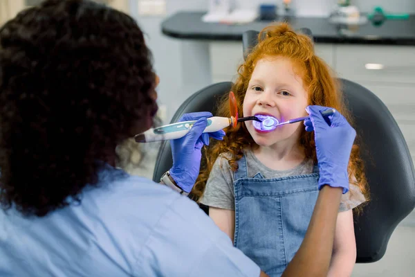 Közelkép a kis aranyos göndör vörös hajú lány nyitott szájjal a kezelés alatt a fogorvosnál. Nő afro-amerikai fogorvos latex kesztyűben, hogy a fogak kitöltése UV lámpa — Stock Fotó