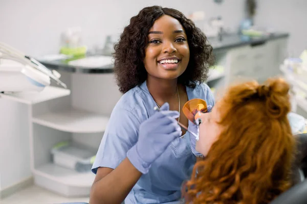 Sorridente dentista donna nera indossa uniforme blu, fornendo restauro dei denti e riempimento con polimerizzazione polimerizzazione lampada UV per il suo piccolo paziente, ragazza carina con i capelli rossi ricci — Foto Stock