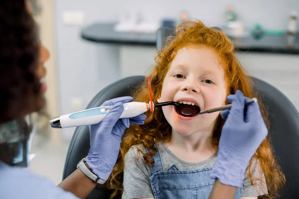 Zamknij widok na małą rudowłosą pacjentkę, która ma leczenie stomatologiczne w gabinecie dentystycznym. Młoda afrykańska dentystka wypełnia zęby polimeryzacją utwardzaną lampą UV — Zdjęcie stockowe