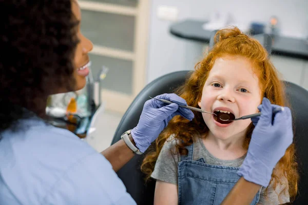 Dziecko w klinice dentystycznej. Zbliżenie portret dziewczynki z czerwonymi kręconymi włosami, siedzącej z otwartymi ustami w fotelu dentystycznym, podczas gdy afrykańska dentystka sprawdza zęby za pomocą lustra dentystycznego. — Zdjęcie stockowe