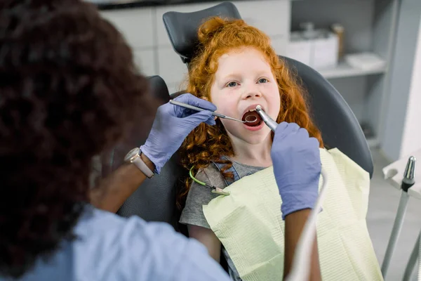 Linda niña de pelo rojo bastante poco durante el procedimiento dental en la clínica dental de los niños modernos. Mujer dentista africana curando los dientes de una niña, utilizando taladro dental y espejo —  Fotos de Stock