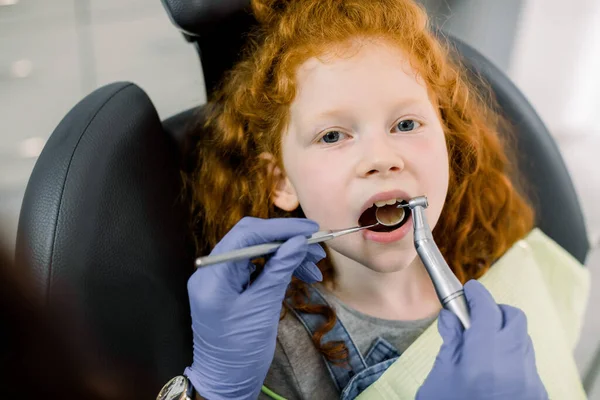 Detailní záběr malé roztomilé kudrnaté zrzavé dívky s otevřenými ústy během ústní prohlídky u zubaře. Ruce zubaře v latexových rukavicích, zpracování zubů vrtačkou a zrcadlem — Stock fotografie
