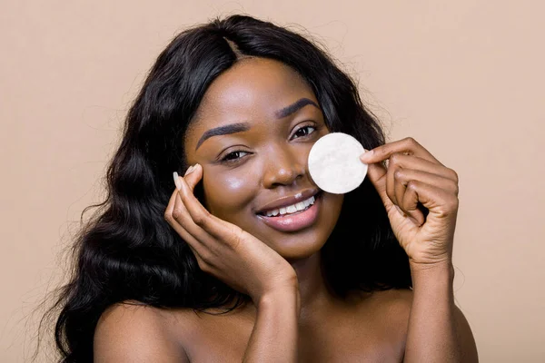 Natural beauty, people, races concept. Close up portrait of pretty smiling young african woman with bare shoulders, cleaning her face with cotton pad, posing over beige background — Fotografia de Stock