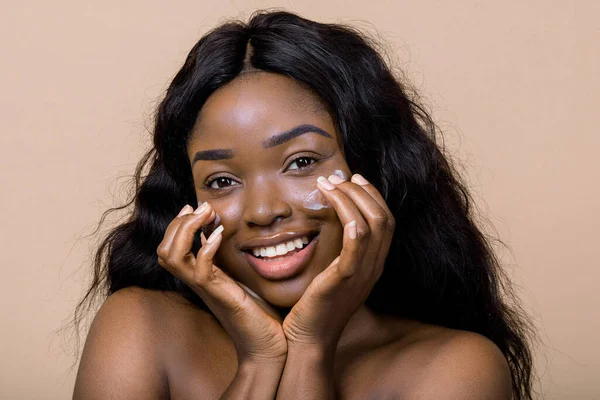 Close up shot of smiling attractive naked dark skinned woman with long hair, appiles face cream on cheeks, poses against studio beige background. Skin health, natural beauty, cosmetic concept — Zdjęcie stockowe