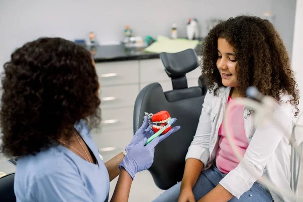 Female African doctor demonstrating proper dental care to her little kid patient, mulatto mixed raced schoolgirl. Happy kid listening and having fun at modern dentists office — Fotografia de Stock