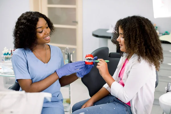 Teeth hygiene. Cute happy child girl with red curly hair, having fun with her mom at dentist office, while trying to brush teeth on jaw model under the supervision of female black dentist — Foto Stock