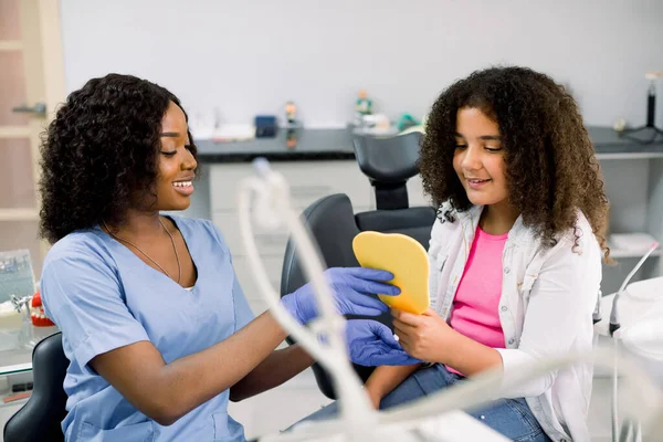 Cute happy mixed raced teen girl, looking at the mirror with toothy smile, sitting on the dental chair while smiling black female dentist showing the result of treatment teeth at the dental office — Fotografia de Stock