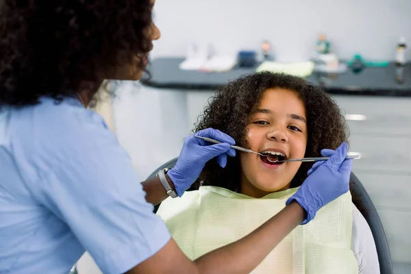 Close up top view of pretty kid, african american curly girl in dentist chair, with mouth open, having dental examination. Back view of female African dentist in gloves, holding tools — Fotografia de Stock