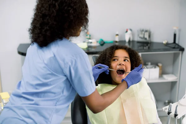 Close up of pretty mulatto mixed raced teen girl in dentist chair, with mouth open, having dental examination. Back view of female African dentist in gloves, holding dental tools — Stock Fotó