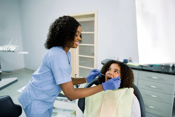 Dentist and kid at modern dental clinic. Young smiling african american dentist in uniform and gloves, making examination of her small patient, little mixed raced curly girl, to prevent caries — Zdjęcie stockowe