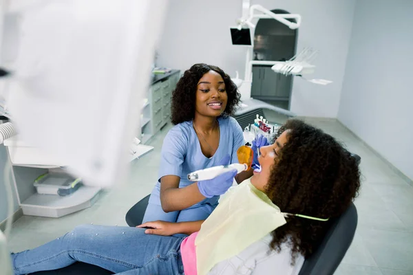 Smiling black woman dentist wearing blue uniform, providing tooth restoration and filling with curing polymerization UV lamp for her little patient, cute african american girl with curly hair