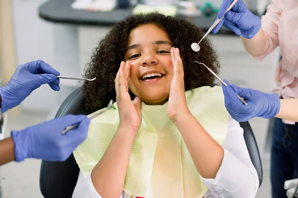 Healthy teeth, caries prevention and pediatric dentistry. Close up of scared funny little mixed raced mulatto girl, looking at camera and screaming and hands of two dentists in gloves with tools — Zdjęcie stockowe