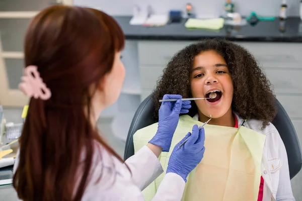 Close up view of pretty kid, african american curly girl in dentist chair, with mouth open, having dental examination. Back view of female dentist in gloves, holding tools — Zdjęcie stockowe