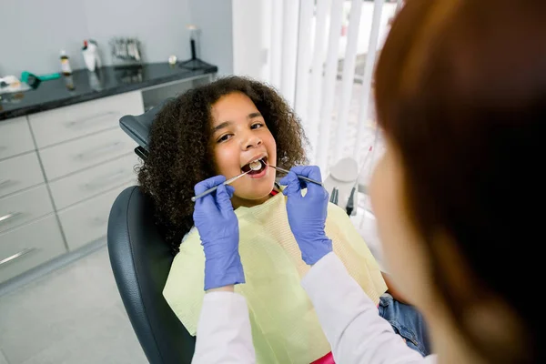 Close-up of cute curly mixed raced girl with open mouth during oral checkup at the dentist. Young female Caucasian dentist in latex gloves, making teeth examination with tools —  Fotos de Stock